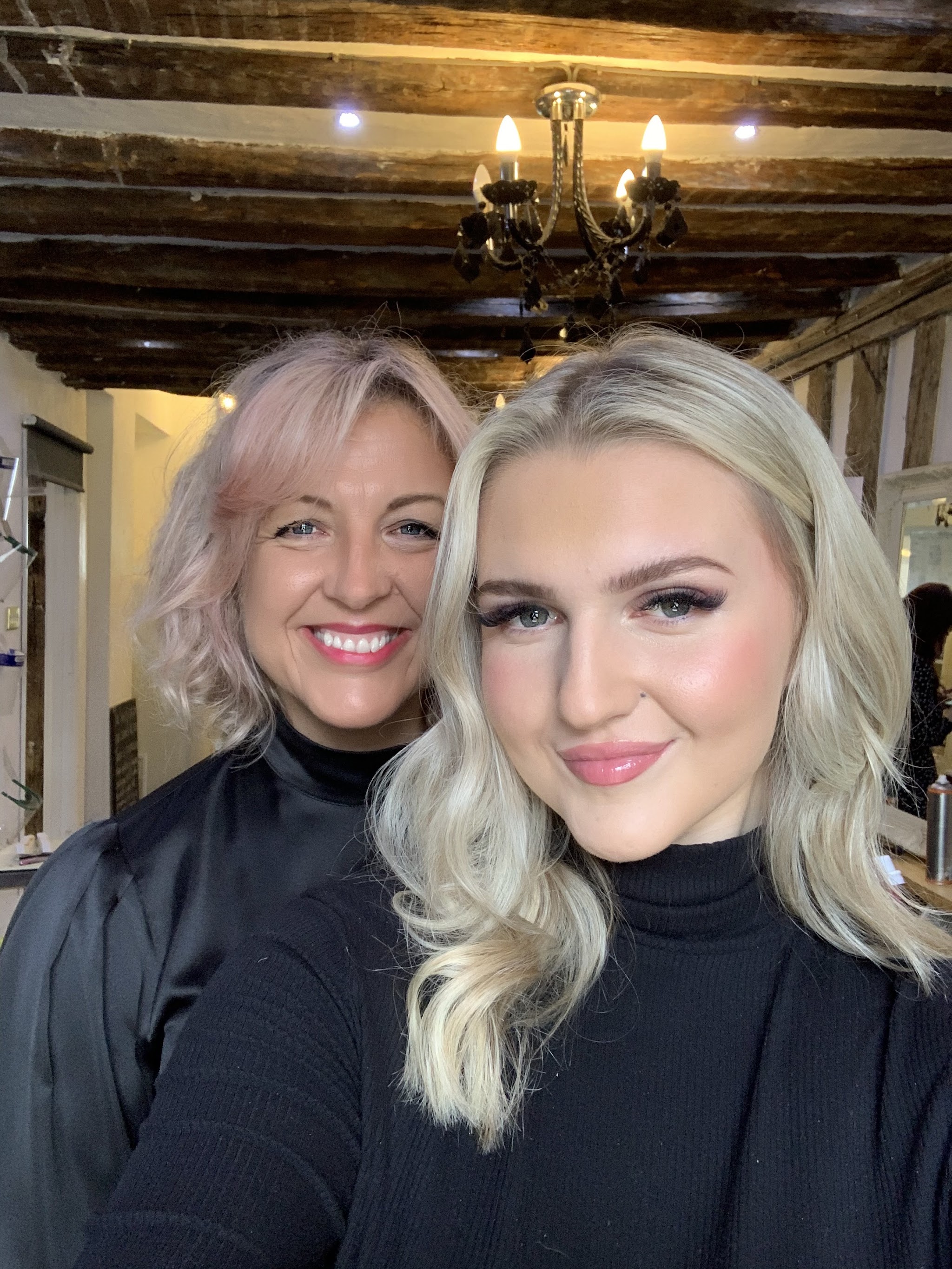 A happy client with platinum curls and her stylist, smiling for a selfie in the beamed salon